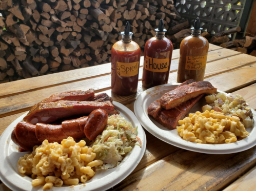 Two plates of barbecue with sausage, ribs, coleslaw, and macaroni, alongside three bottles of sauce labeled "Spicy," "House," and "Sweet."