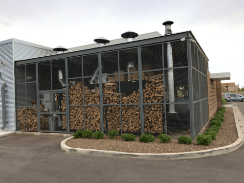 A fenced storage area filled with neatly stacked firewood beside a building, with ventilation pipes visible above.
