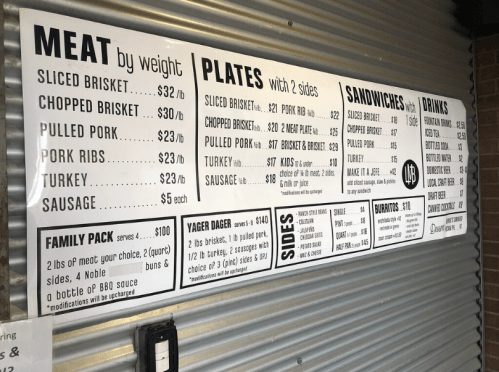 Menu board displaying prices for meat, plates, sandwiches, drinks, and sides at a barbecue restaurant.