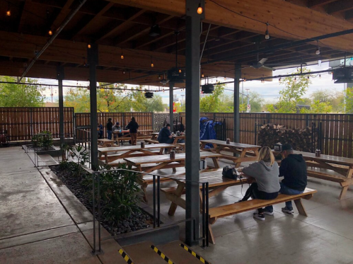A spacious outdoor dining area with wooden tables, plants, and string lights, mostly empty with a few people seated.