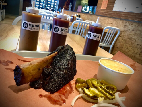 A plate with a large smoked beef rib, pickles, onions, jalapeños, and a side of creamy sauce, with sauce bottles in the background.
