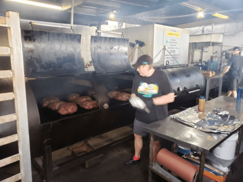 A chef tends to large cuts of meat on a smoker in a busy kitchen, with smoke rising and another worker in the background.