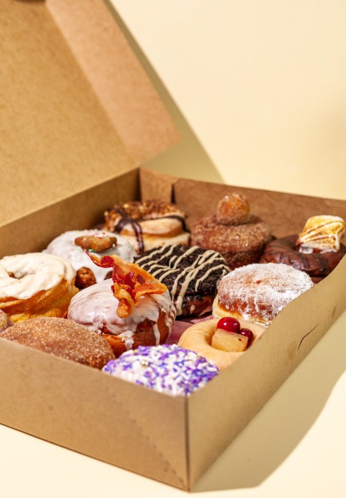 A variety of colorful donuts in a brown box, featuring toppings like bacon, chocolate, and sprinkles.