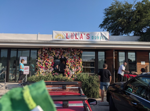 A vibrant storefront of Lola's Donuts adorned with flowers, with people entering and enjoying the sunny day.