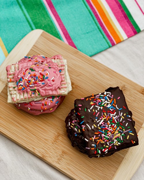 A wooden board with a frosted pastry and a chocolate-covered treat, both topped with colorful sprinkles.