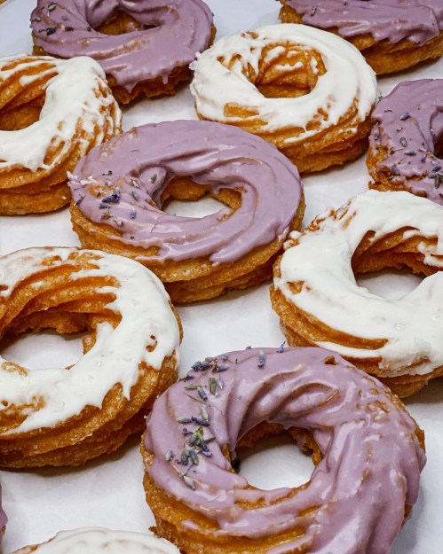 A close-up of colorful donuts with lavender and white icing, some topped with lavender sprinkles.