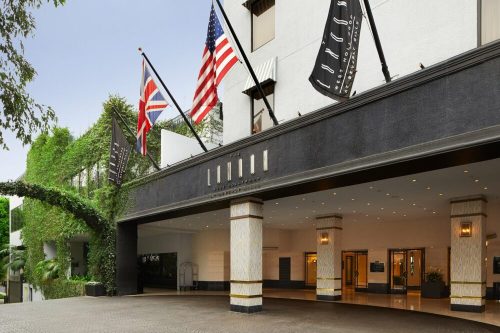 Entrance of a hotel with flags, including the American and British flags, surrounded by greenery.