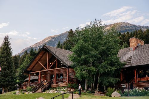 A rustic log cabin surrounded by trees and mountains, with a person standing near the entrance.