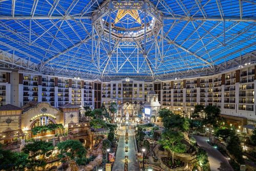 A spacious indoor atrium with a glass ceiling, featuring lush greenery, water features, and hotel rooms in the background.