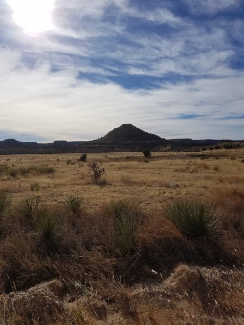 A vast, dry landscape with sparse vegetation and a distant hill under a bright sky with scattered clouds.