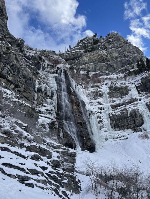 A frozen waterfall cascading down rocky cliffs, surrounded by snow and icy formations under a blue sky.