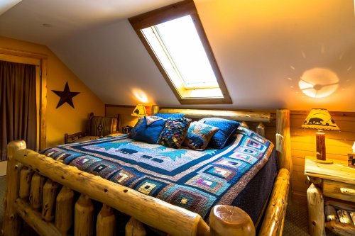 Cozy attic bedroom with a skylight, log furniture, and a colorful quilt on a large bed. Warm, inviting atmosphere.