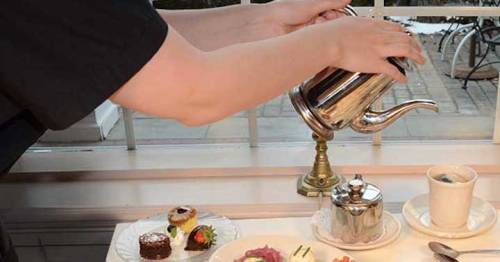 A person pours tea from a silver teapot over a table set with pastries and a cup.