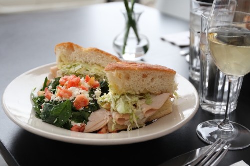 A plate with a sandwich filled with turkey and lettuce, accompanied by a fresh salad and a glass of white wine.