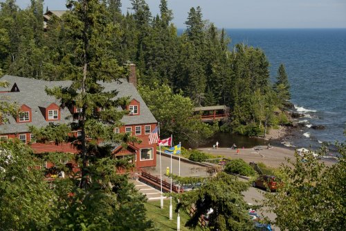 A scenic view of a lodge by the water, surrounded by trees, with flags and a beach in the background.