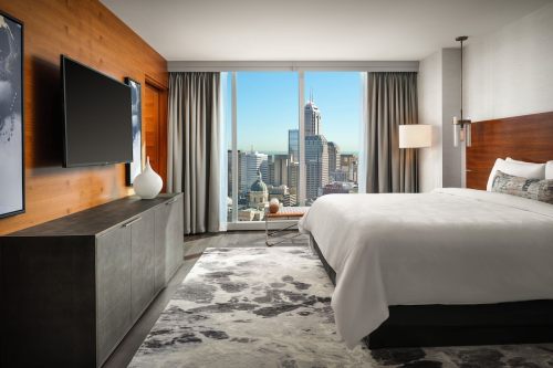 Modern hotel room with a large window showcasing a city skyline, featuring a bed, TV, and stylish decor.