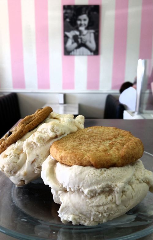 Two ice cream sandwiches with cookie layers, displayed on a glass plate in a pink-striped ice cream shop.