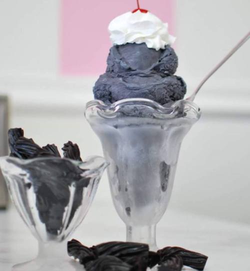 A glass dish filled with dark gray ice cream topped with whipped cream and a cherry, alongside a bowl of black licorice.