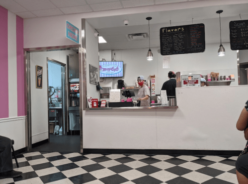 A small ice cream shop interior with a counter, menu board, and staff serving customers. Black and white checkered floor.
