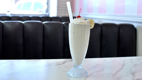 A tall glass of milkshake topped with whipped cream, a cherry, and a slice of banana, set on a marble table.