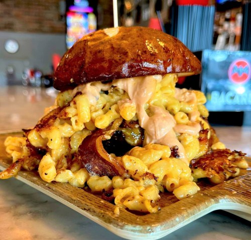 A pretzel bun filled with creamy macaroni and cheese, topped with sauce, served on a wooden plate.