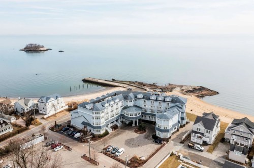 Aerial view of a coastal hotel near a sandy beach and calm water, with a small island in the distance.