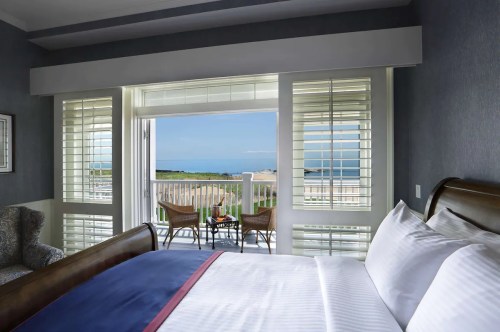 A cozy hotel room with a bed, overlooking a balcony with chairs and a view of the ocean through open shutters.