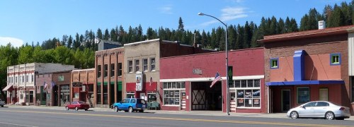 A small town street lined with colorful storefronts and parked cars, surrounded by trees under a clear blue sky.