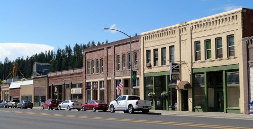 Historic buildings line a street, featuring shops and an American flag, with trees and a blue sky in the background.