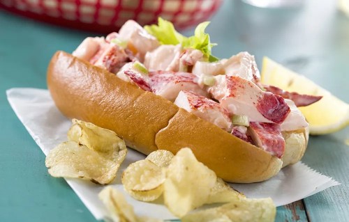 A lobster roll filled with lobster meat and lettuce, served with potato chips and a lemon wedge on the side.