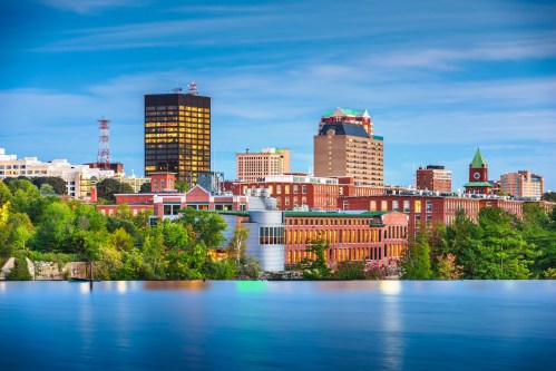 City skyline featuring modern and historic buildings along a river, with greenery in the foreground and a clear blue sky.