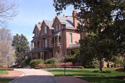A large, elegant brick house with multiple gables, surrounded by lush greenery and colorful flowers on a sunny day.