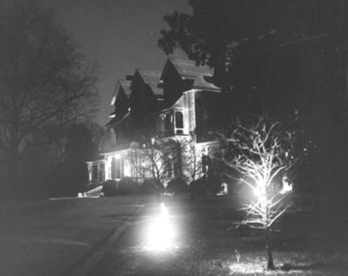 A dark, eerie house illuminated by lights, with a small tree glowing in the foreground.