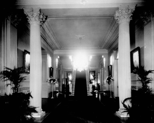 A grand hallway with tall columns, a chandelier, and a staircase, adorned with plants and framed artwork.