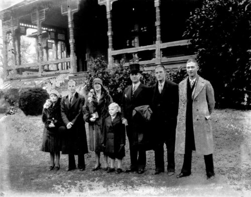 A black-and-white photo of a group of seven people standing outside a large wooden building, dressed in early 20th-century attire.