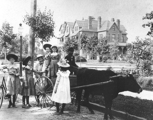 A group of children in vintage clothing poses with a cow and a cart in front of a large house and trees.
