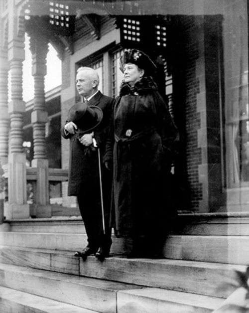 A man and woman in formal attire stand on steps, with ornate architecture in the background, captured in black and white.