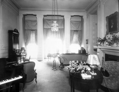 A vintage living room with elegant furnishings, large windows, and a piano, illuminated by natural light.