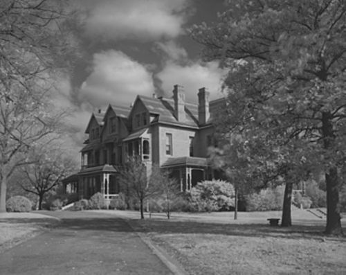 A large, historic house surrounded by trees and shrubs, set against a cloudy sky in black and white.
