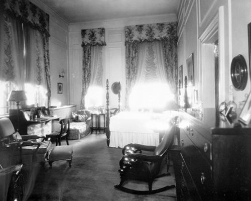 A vintage bedroom with ornate curtains, a large bed, a rocking chair, and antique furniture in soft lighting.