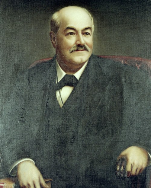 Portrait of a middle-aged man with a mustache, wearing a dark suit and seated in a chair, smiling gently.