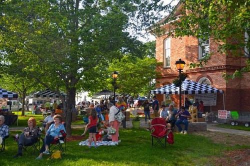 A lively outdoor market scene with people sitting on grass, tents, and a historic brick building in the background.