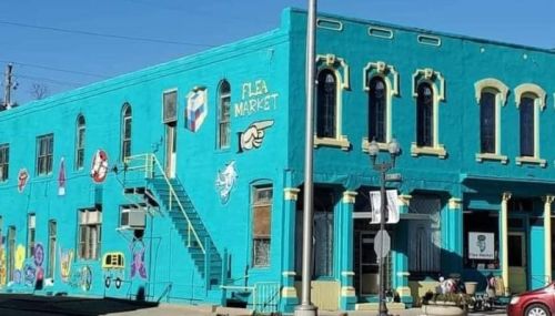 Bright turquoise building with murals, featuring a sign for a flea market and an outdoor staircase.