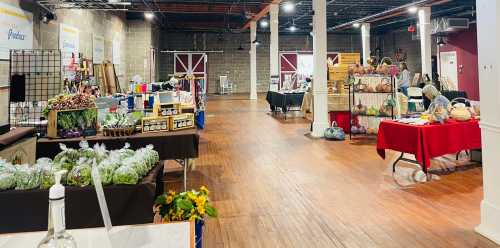 A spacious indoor market with vendor tables displaying crafts, produce, and handmade goods in a well-lit setting.