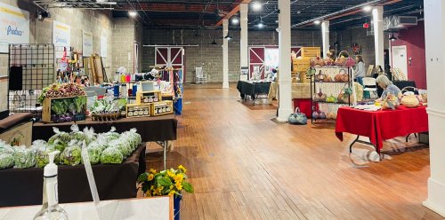 A spacious indoor market with vendor tables displaying crafts, produce, and handmade goods in a well-lit setting.