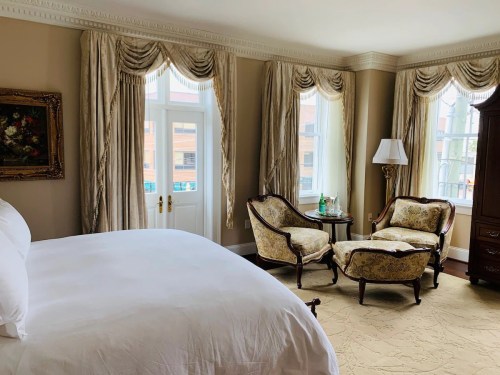 Elegant hotel room featuring a bed, two armchairs, a lamp, and large windows with drapes.