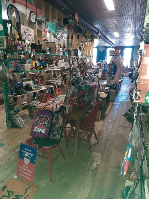 A cluttered vintage shop interior with various items for sale, including backpacks and furniture, under warm lighting.