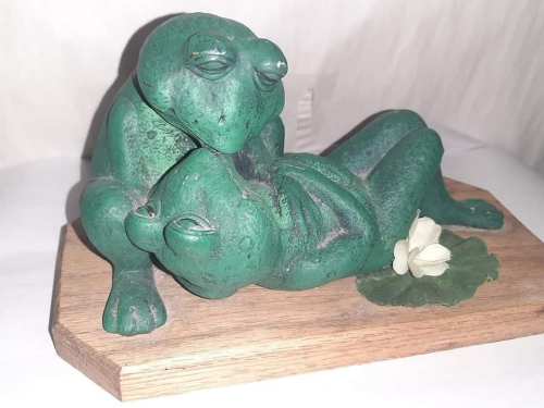 Two green frog sculptures in a playful pose on a wooden base, with a small flower and leaf nearby.