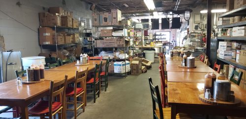 A rustic dining area with wooden tables and chairs, surrounded by shelves of supplies and boxes in a warehouse setting.