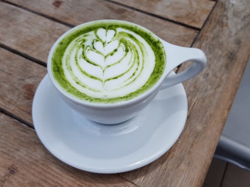 A cup of matcha latte with intricate leaf latte art, sitting on a white saucer on a wooden table.